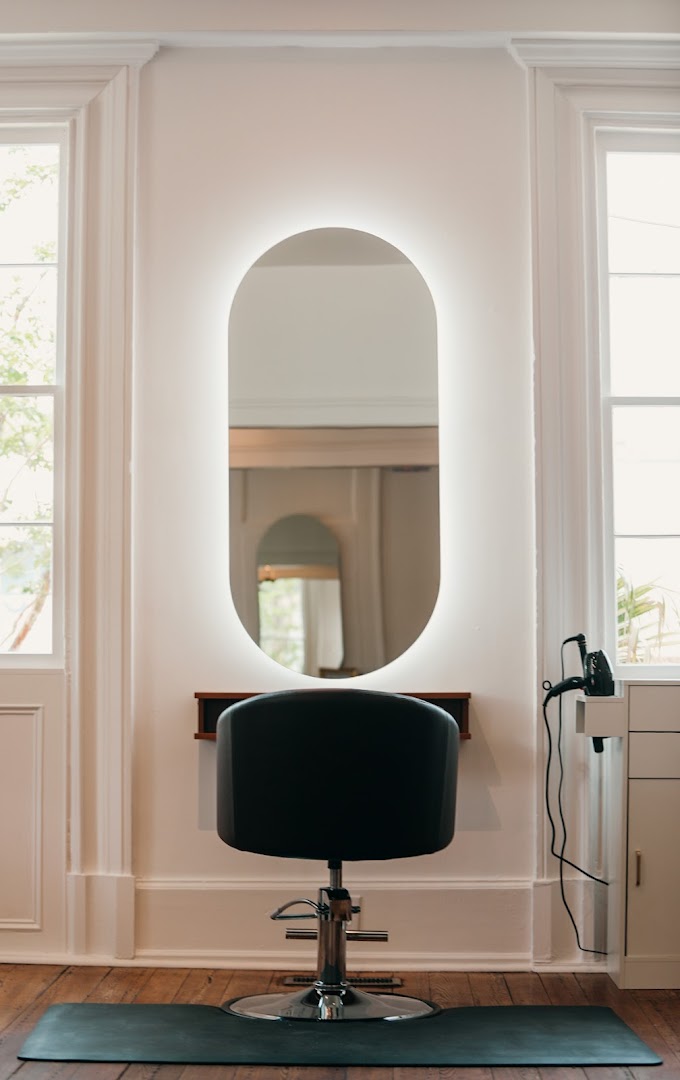 Inside Common Citizen — a black salon chair below an oval LED-lit mirror, on wood plank floors, between two windows letting in tree-foliage light.