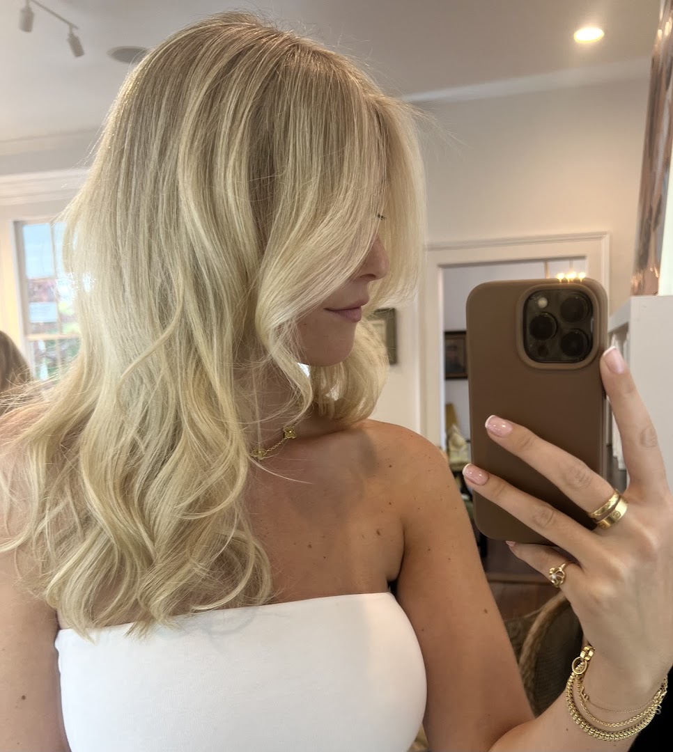 A client in a mirror-selfie post-blowout at Common Citizen — long blonde waves, white tube top, hand holding iPhone.