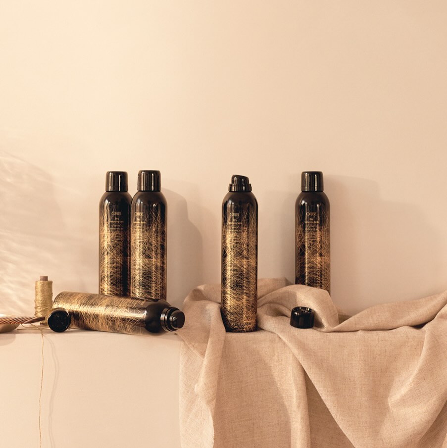A still-life of Oribe Dry Texturizing Spray bottles in their bronze pattern, arranged on cream linen — Common Citizen product editorial.